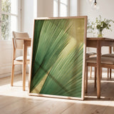 a large framed print of a green leaf with a yellow center, displayed on a wooden floor in a room with a dining table and chairs.