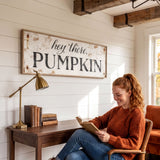 A woman is sitting at a desk, reading a book, with a lamp and a stack of books nearby. Above her, there is a sign that says "hey there, PUMPKIN".