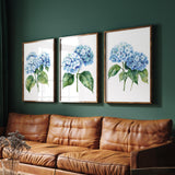 a cozy living room with a brown leather couch and a wooden coffee table. Above the couch, there are three framed watercolor paintings of blue hydrangeas hanging on the wall.