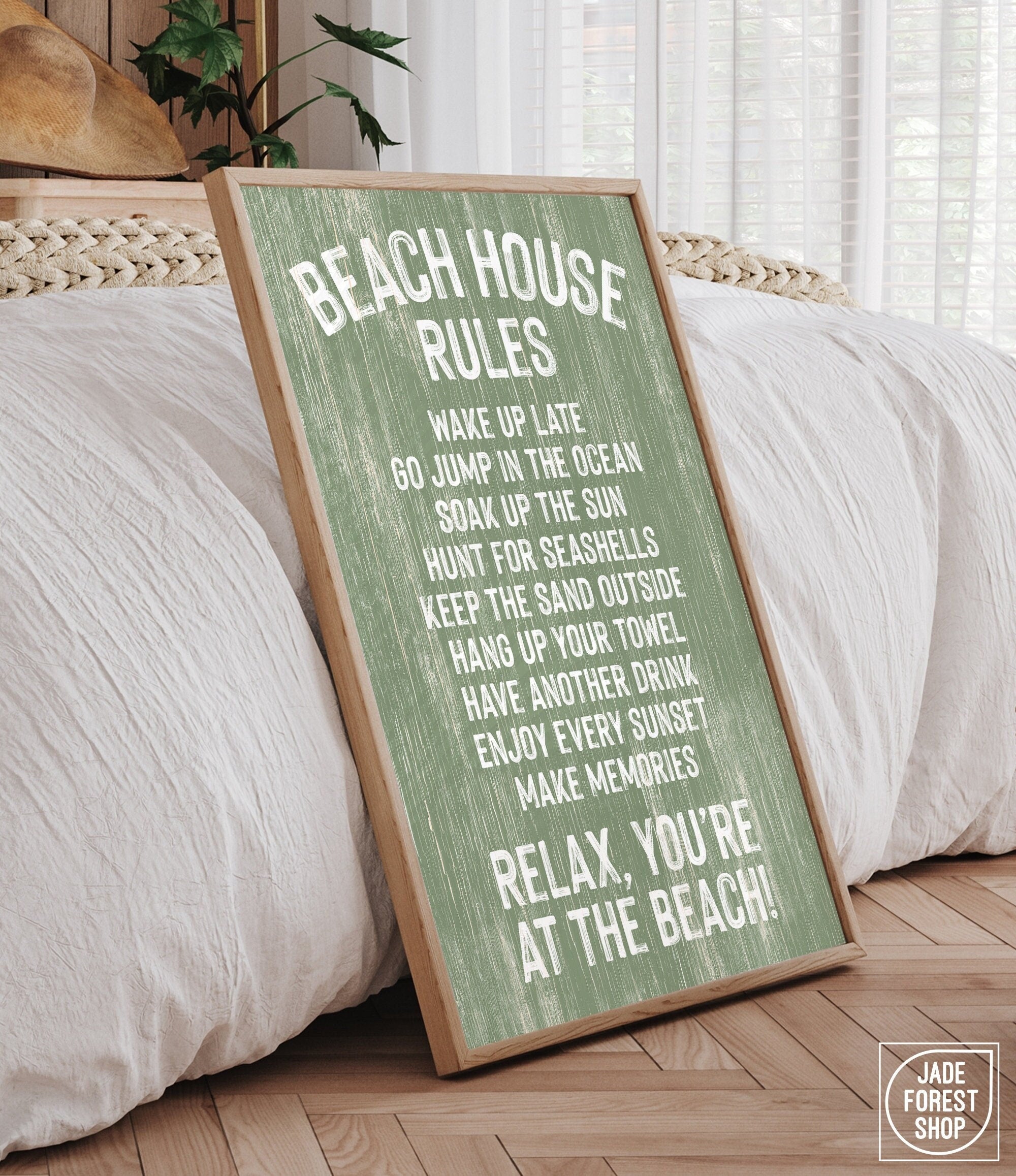 The Beach House Rules Sign, Painted Wood, Seagrass, with white text, leans against a bed in a sunlit bedroom, bringing cozy coastal charm and stylish wall art to the space.