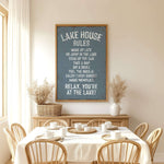 a dining room with a table set for a meal, surrounded by wooden chairs. Above the table, there is a framed sign with a list of rules for the lake house, including instructions for guests to relax, enjoy the sunset, and make memories.