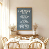 a dining room with a table set for a meal, surrounded by wooden chairs. Above the table, there is a framed sign with a list of rules for the lake house, including instructions for guests to relax, enjoy the sunset, and make memories.