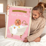A woman is sitting on a bed holding a framed poster with a cocker spaniel dog in a bathtub.