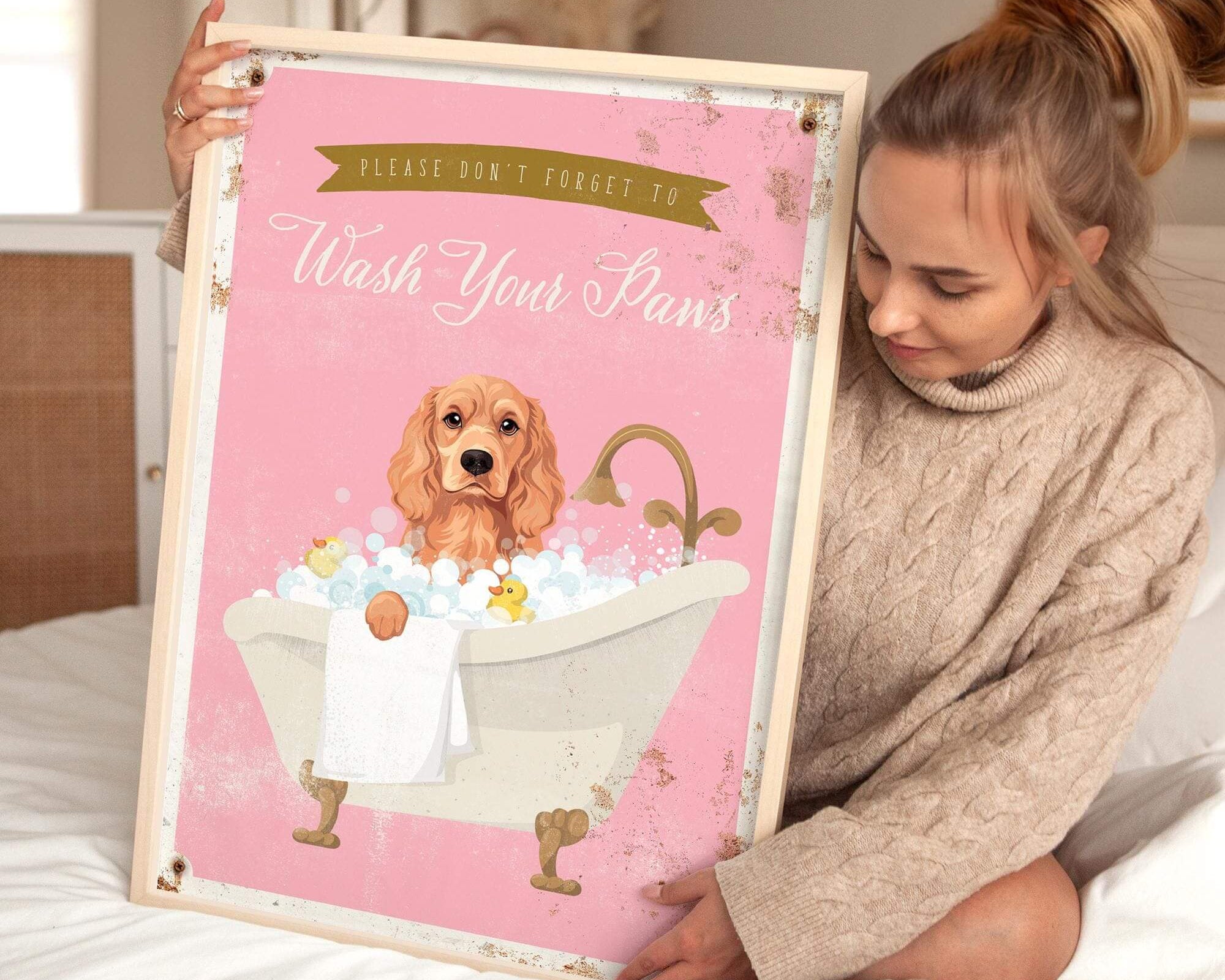 A woman is sitting on a bed holding a framed poster with a cocker spaniel dog in a bathtub.