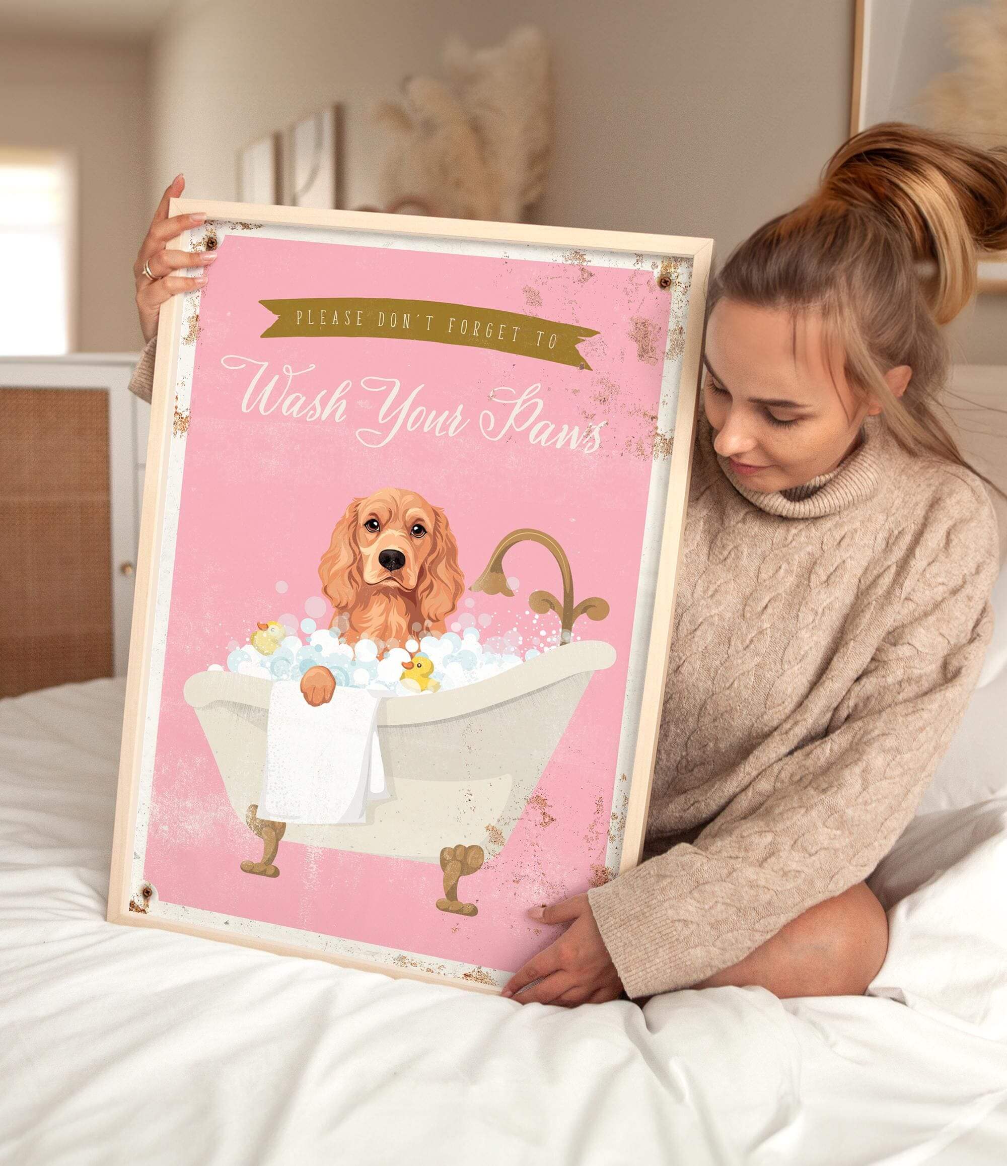 A woman is sitting on a bed holding a framed poster with a cocker spaniel dog in a bathtub.