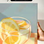 a close-up of a glass containing a slice of orange, with a hand holding the canvas.
