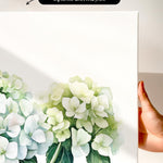a hand holding a white canvas print with a watercolor painting of white hydrangea flowers.