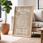 A wooden sign with white text that reads "BEACH HOUSE RULES" is displayed in a living room, with a potted plant and a couch visible in the background.