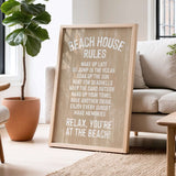 A wooden sign with white text that reads "BEACH HOUSE RULES" is displayed in a living room, with a potted plant and a couch visible in the background.