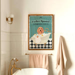 a framed poster of a cocker spaniel in a bathtub, with a towel and towel rack visible in the foreground.