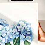 a hand holding a white canvas print of a watercolor painting of blue hydrangea flowers.