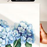 a hand holding a white canvas print of a watercolor painting of blue hydrangea flowers.