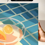 a hand holding a framed print of a colorful abstract design with a glass containing orange slices.