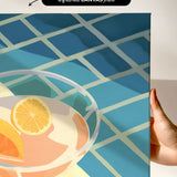 a hand holding a framed print of a colorful abstract design with a glass containing orange slices.