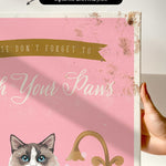 a hand holding a framed canvas print with a cat illustration and text.