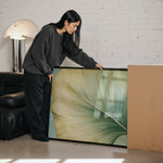 A woman in a gray sweater is leaning over a large framed print, examining it with interest.
