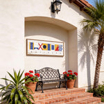a white building with a black wrought iron bench and two potted plants, one of which has red flowers. Above the bench, there is a framed sign with colorful nautical symbols.