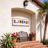 a white building with a black wrought iron bench and two potted plants, one of which has red flowers. Above the bench, there is a framed sign with colorful nautical symbols.