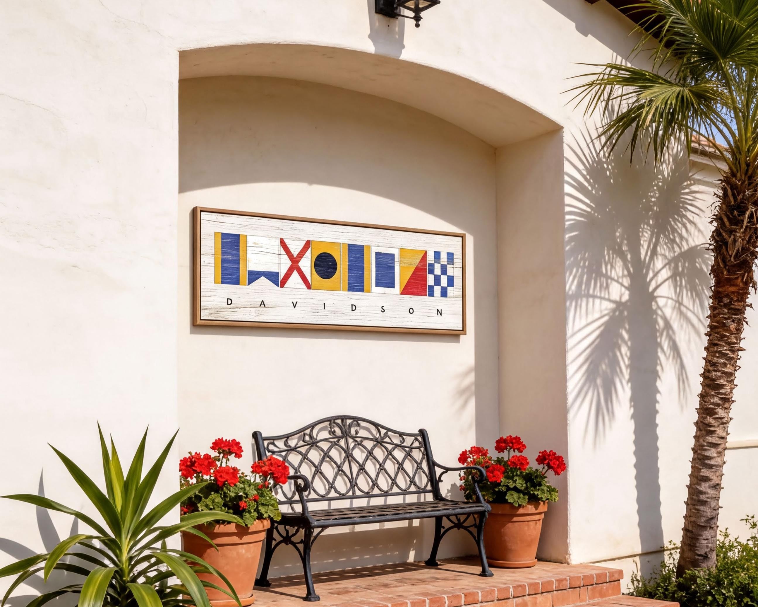 a white building with a black wrought iron bench and two potted plants, one of which has red flowers. Above the bench, there is a framed sign with colorful nautical symbols.