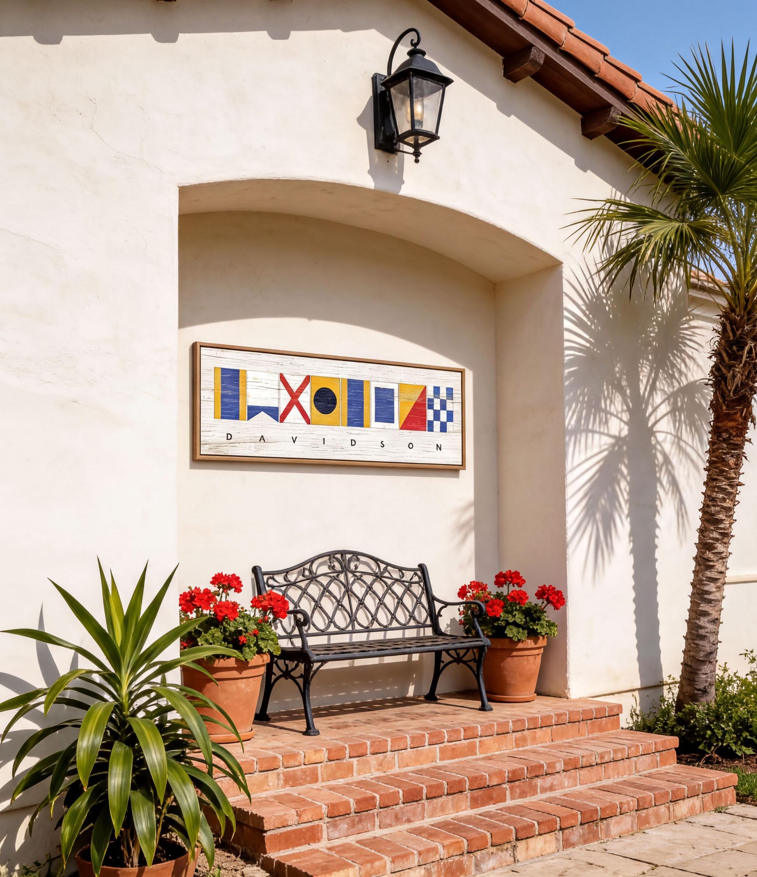 a white building with a black wrought iron bench and two potted plants, one of which has red flowers. Above the bench, there is a framed sign with colorful nautical symbols.