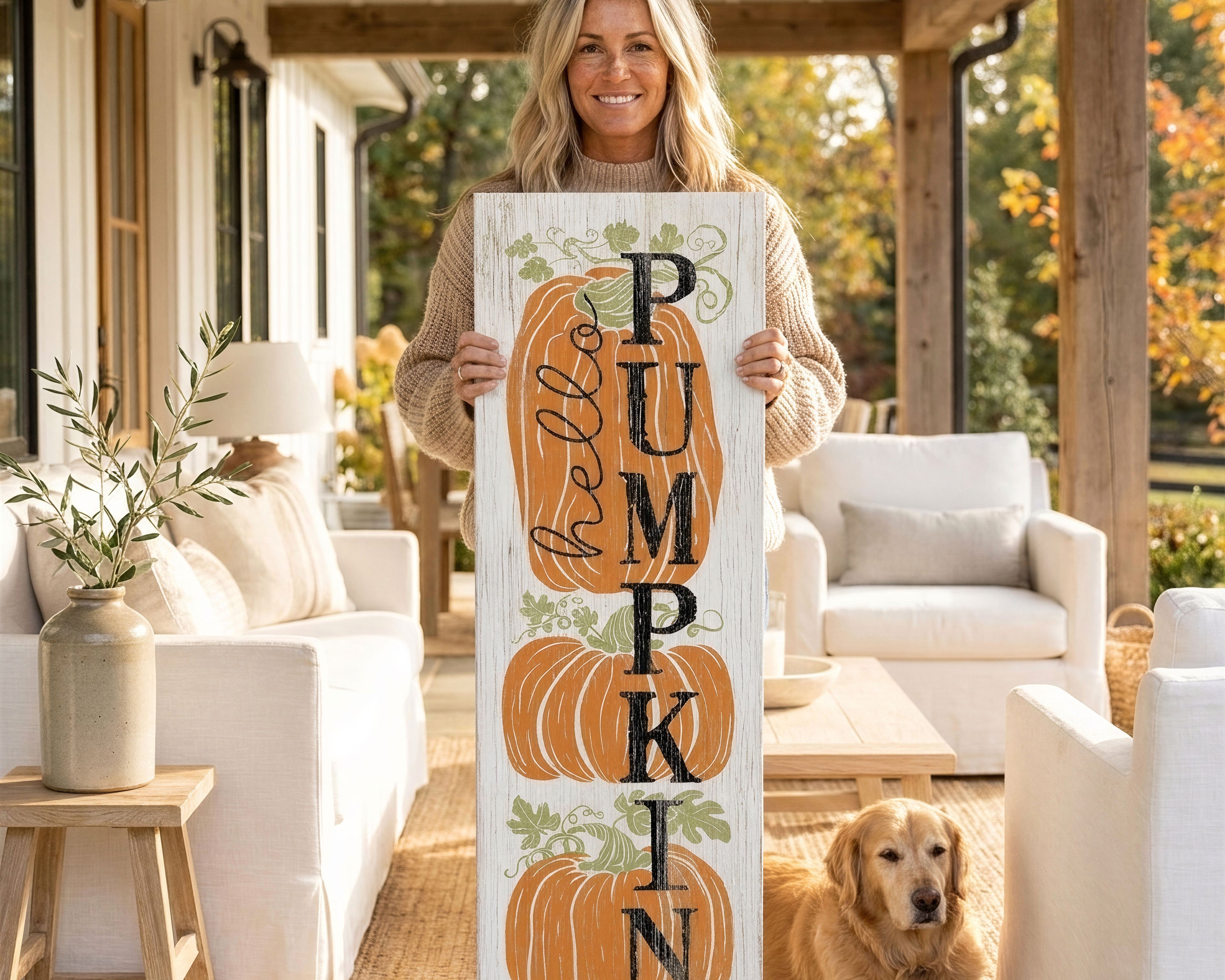 A woman stands on a porch holding a wooden sign with the word "PUMPKIN" written on it, accompanied by a golden retriever dog.