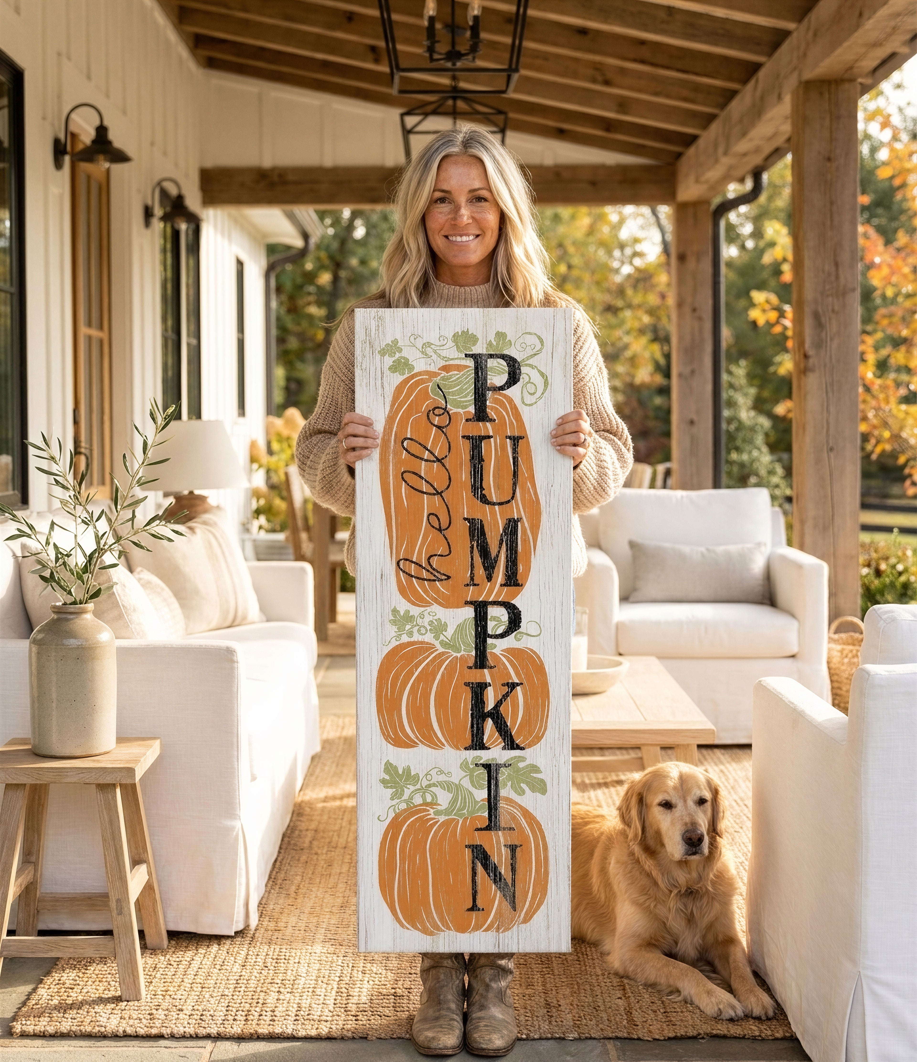 A woman stands on a porch holding a wooden sign with the word "PUMPKIN" written on it, accompanied by a golden retriever dog.