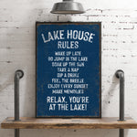 A blue sign with white text that reads "LAKESHOUSE RULES" is displayed on a wooden shelf against a white brick wall.