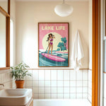 a bathroom with a framed poster of a woman paddleboarding, a potted plant, a sink, and a towel.