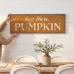 A person is holding up a rustic wooden sign that says "hey there, PUMPKIN" above a dining table with white chairs.