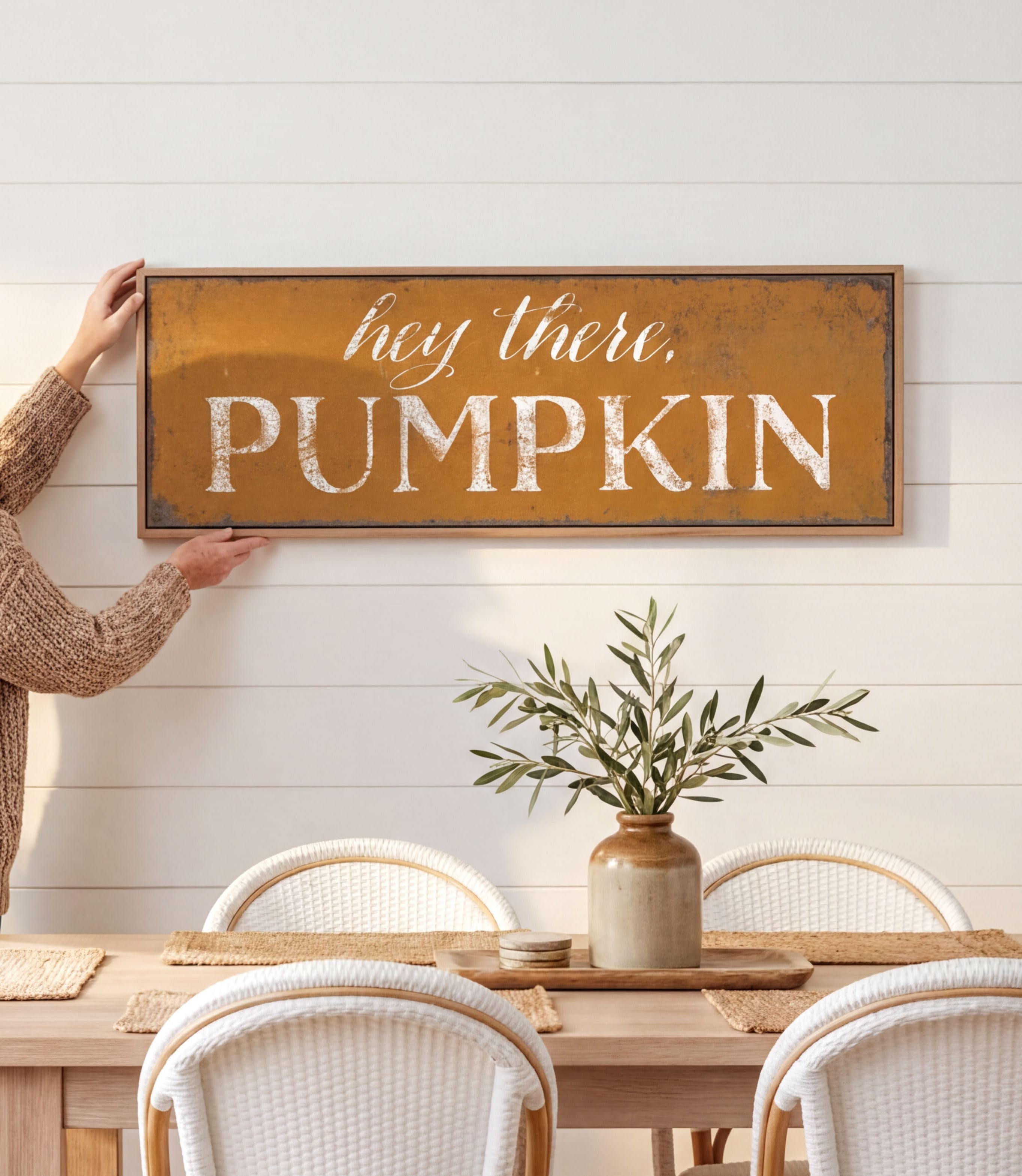 A person is holding up a rustic wooden sign that says "hey there, PUMPKIN" above a dining table with white chairs.