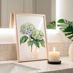 a framed painting of white hydrangeas and green leaves, placed on a bathroom counter next to a lit candle.