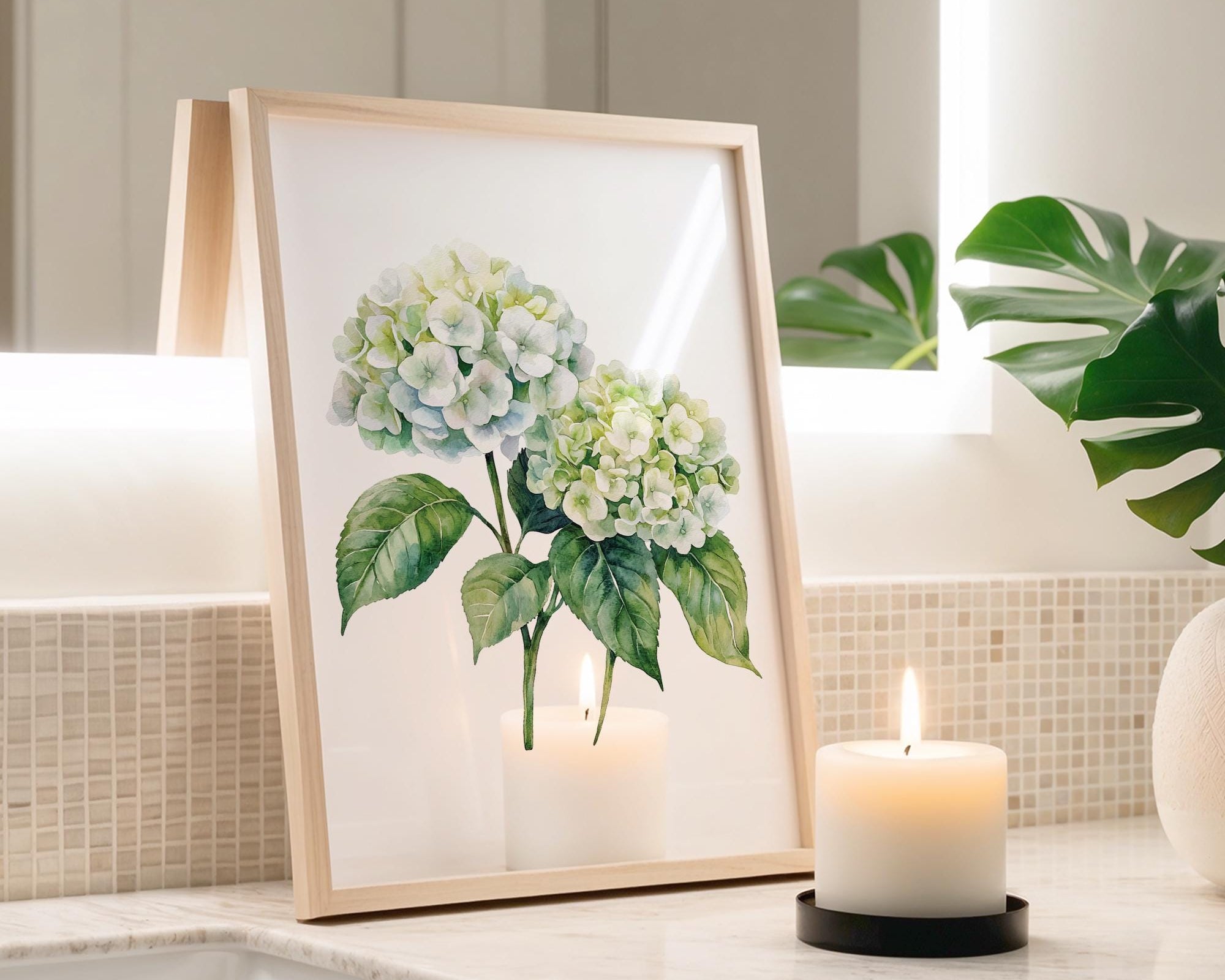 a framed painting of white hydrangeas and green leaves, placed on a bathroom counter next to a lit candle.