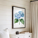 a framed watercolor painting of blue hydrangeas hanging on a white wall above a white dresser with a stack of books on top.