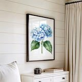 a framed watercolor painting of blue hydrangeas hanging on a white wall above a white dresser with a stack of books on top.