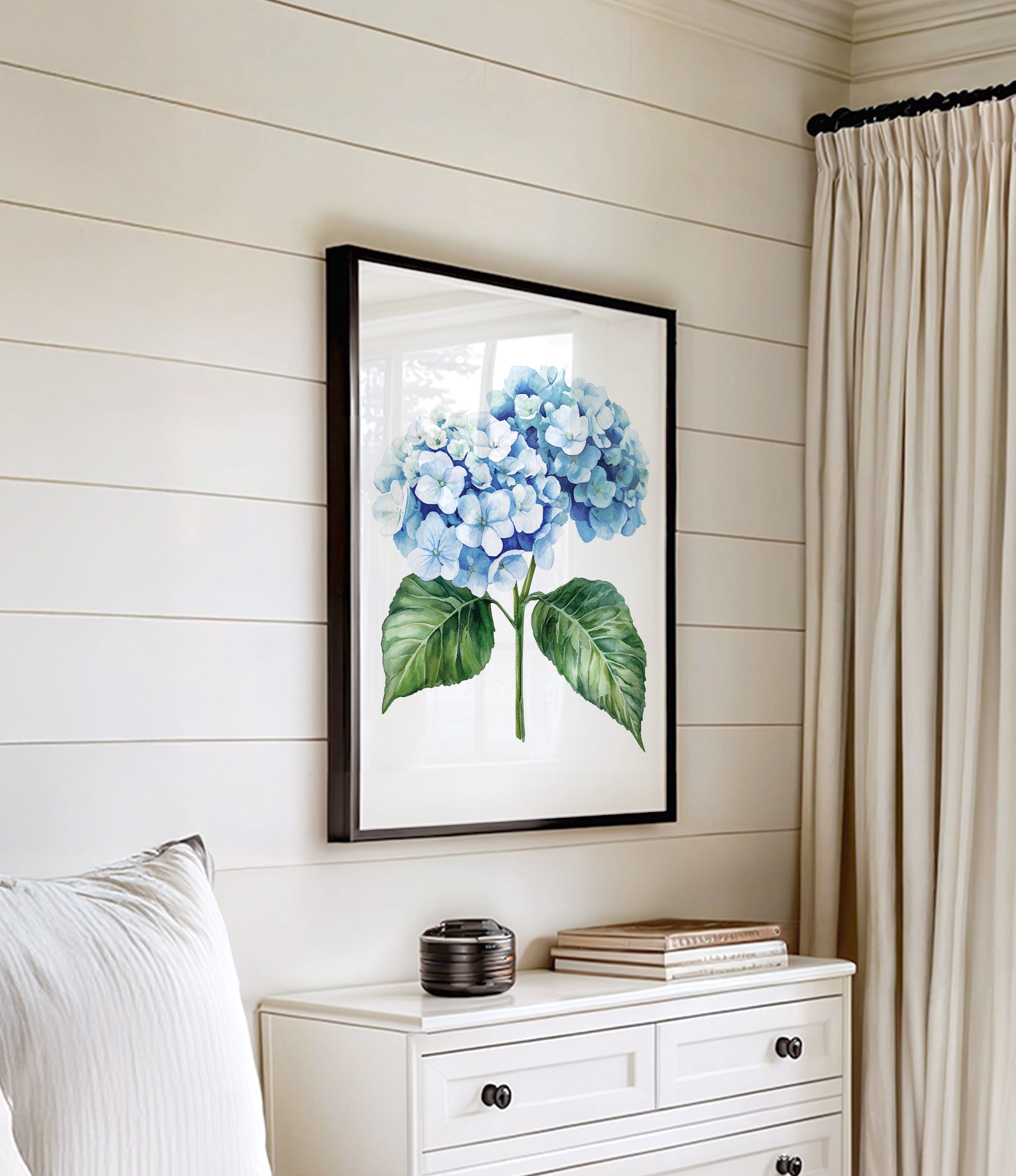a framed watercolor painting of blue hydrangeas hanging on a white wall above a white dresser with a stack of books on top.