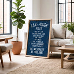 A framed sign on a wooden floor in a living room, displaying a list of rules for a lake house, with a potted plant and a couch in the background.