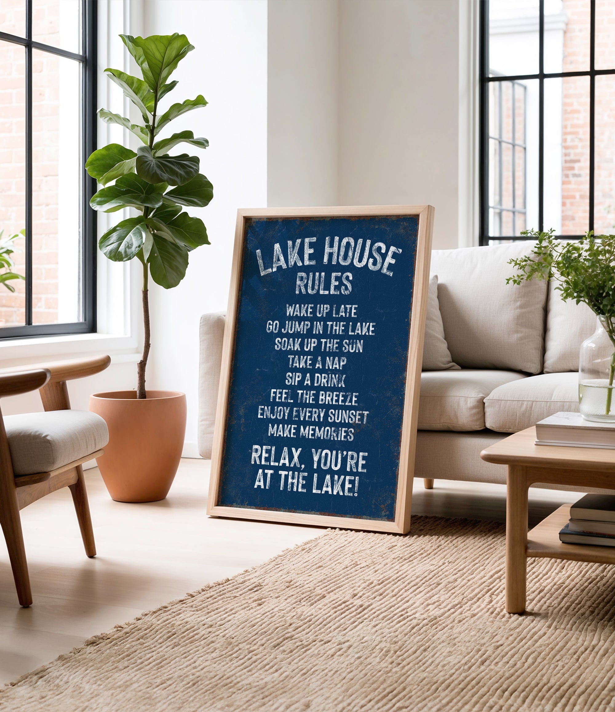 A framed sign on a wooden floor in a living room, displaying a list of rules for a lake house, with a potted plant and a couch in the background.