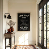 a hallway with a framed sign that reads "LAKESHORE RULES" hanging on the wall. The hallway has a wooden table with a vase of flowers, a rug, and a large window with a view of trees outside.