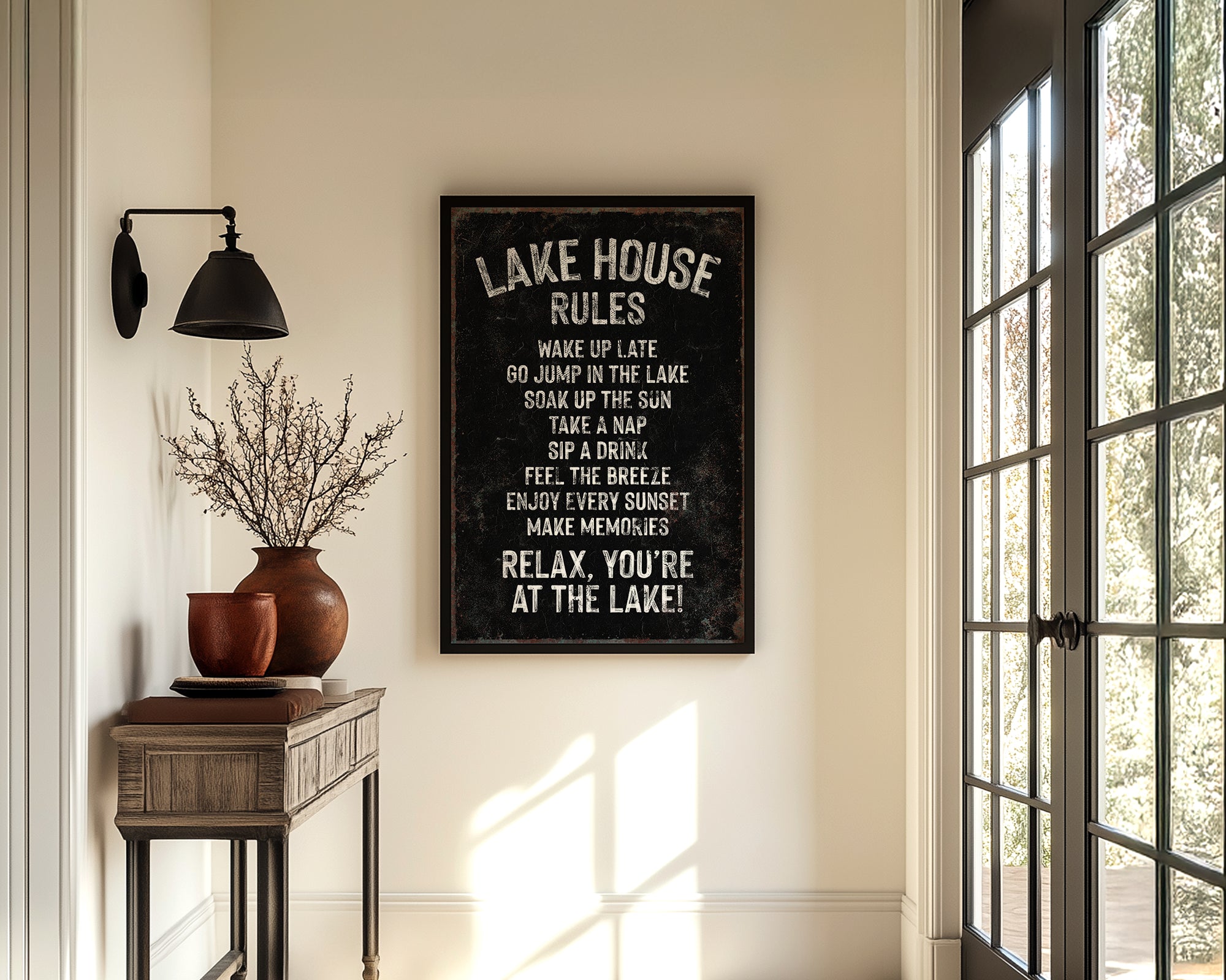 a hallway with a framed sign that reads "LAKESHORE RULES" hanging on the wall. The hallway has a wooden table with a vase of flowers, a rug, and a large window with a view of trees outside.
