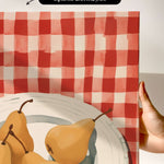 a hand holding a framed print of a red and white checkered tablecloth with a bowl containing three pears.