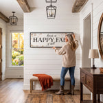 A woman hangs a "Happy Fall" sign on a wall in a hallway with a wooden floor and a window overlooking a garden.