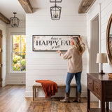 A woman hangs a "Happy Fall" sign on a wall in a hallway with a wooden floor and a window overlooking a garden.