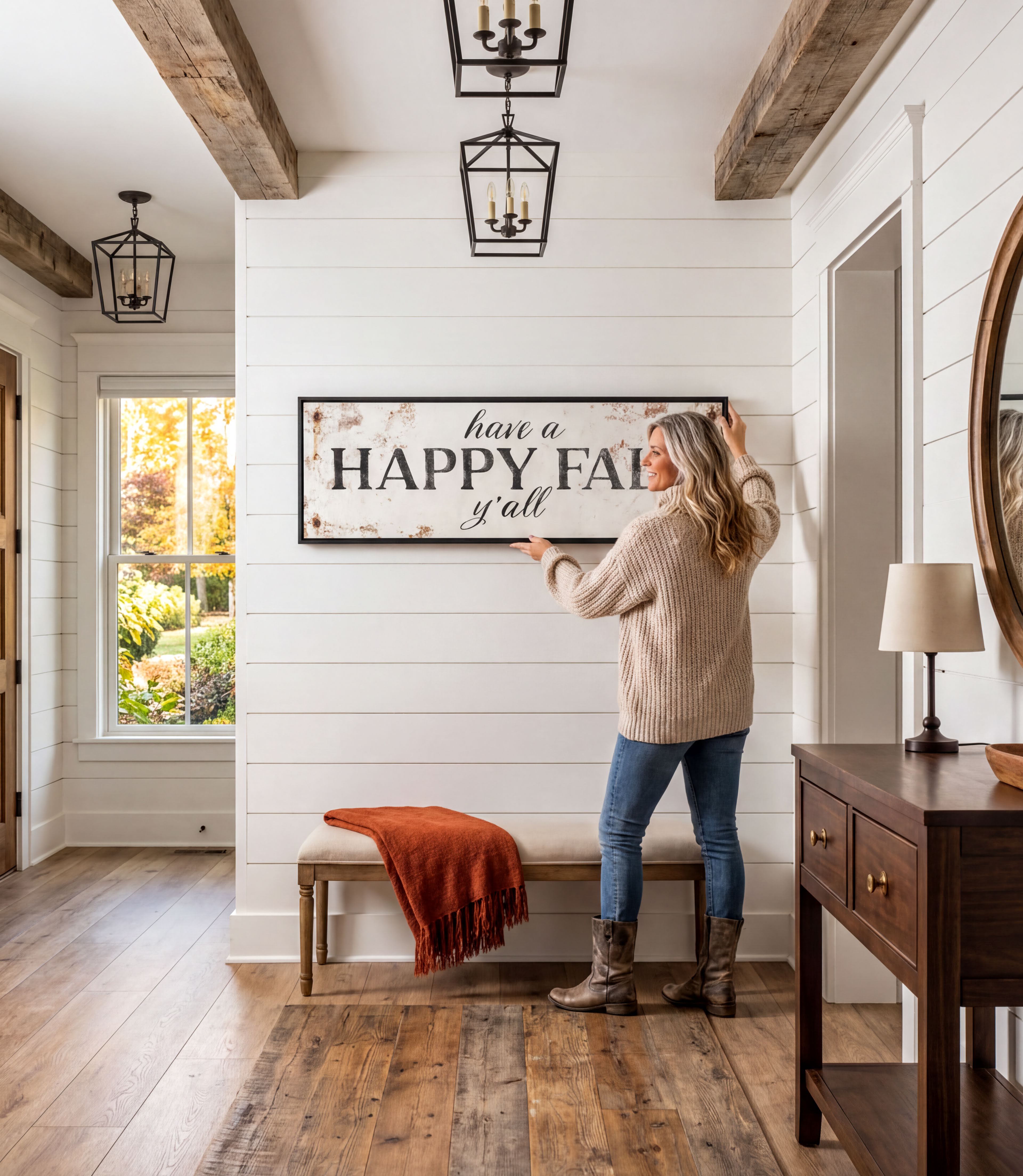 A woman hangs a "Happy Fall" sign on a wall in a hallway with a wooden floor and a window overlooking a garden.