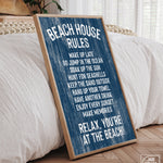 A "Beach House Rules Sign, Painted Wood, Nautical" leans against a bed in a bright, cozy bedroom, bringing charming coastal style and unique wall art to the space.