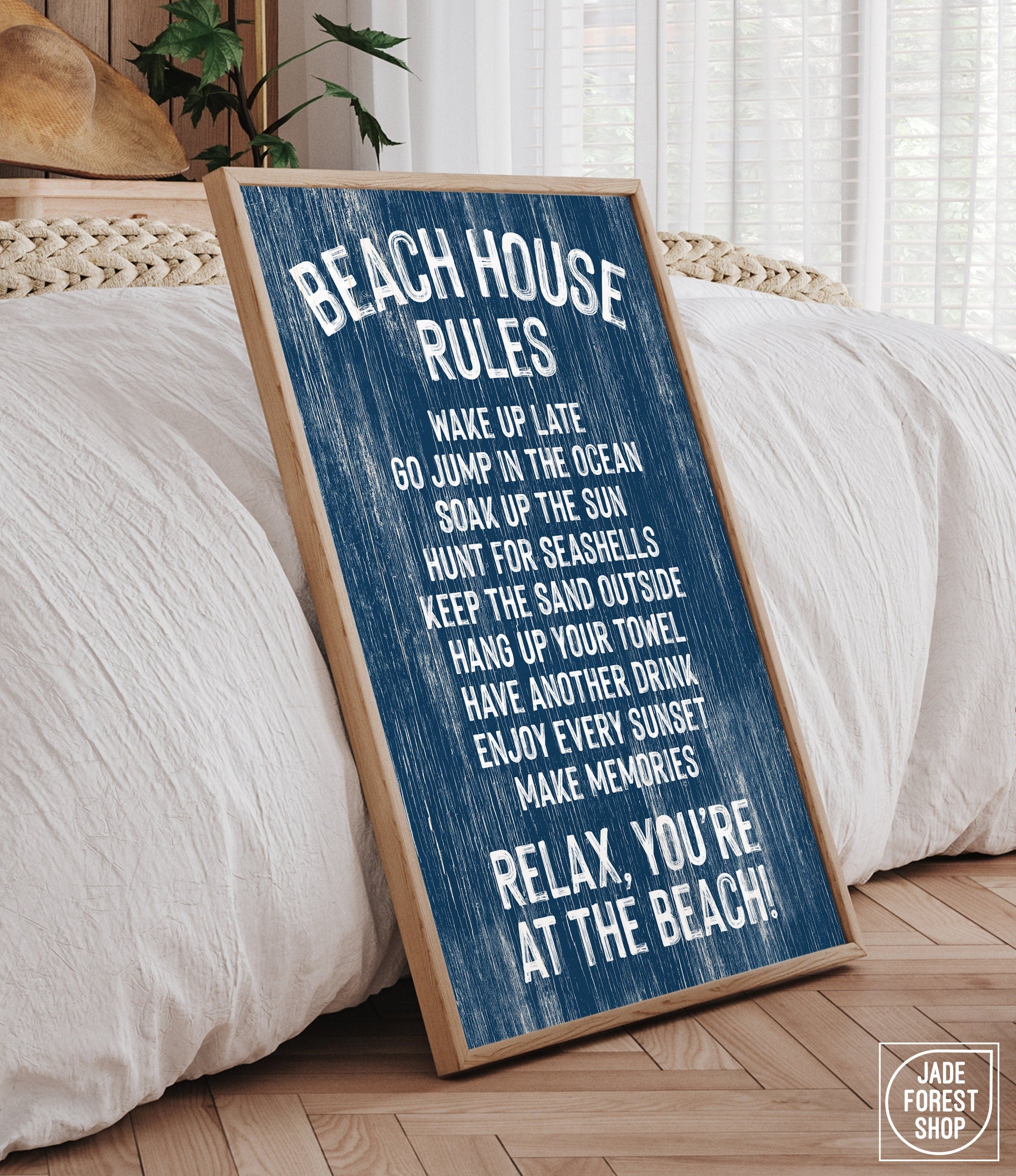 A "Beach House Rules Sign, Painted Wood, Nautical" leans against a bed in a bright, cozy bedroom, bringing charming coastal style and unique wall art to the space.