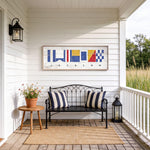 a porch with a bench, a small table, and a lantern, all set against a white wooden wall with a framed nautical flag artwork above it.