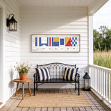 a porch with a bench, a small table, and a lantern, all set against a white wooden wall with a framed nautical flag artwork above it.