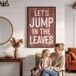 A woman and a young girl are sitting together in a cozy living room, with a large framed sign above them that reads "Let's JUMP IN THE LEAVES".