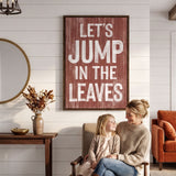 A woman and a young girl are sitting together in a cozy living room, with a large framed sign above them that reads "Let's JUMP IN THE LEAVES".