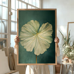 A person is holding up a framed painting of a large, vibrant green leaf against a dark green background.